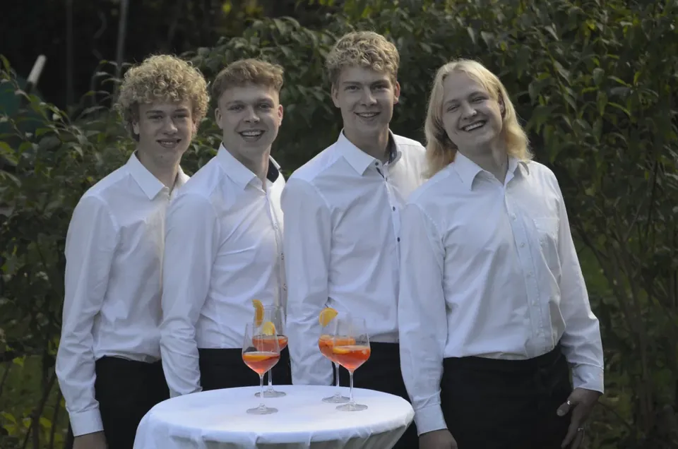 Die vier Brüder stehen lachend nebeneinander, vor ihnen Gläser mit Aperol Spritz