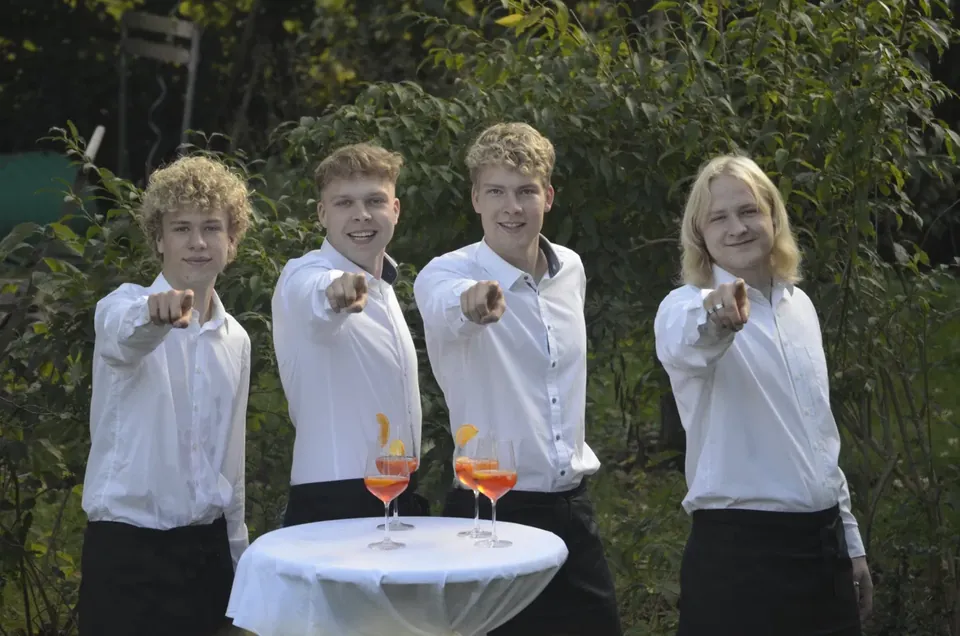 Die vier Brüder in weißen Hemden zeigen lachend in die Kamera, vor ihnen ein Stehtisch mit Aperol Spritz